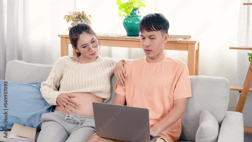 4K, Asian couple. Pregnant wife sitting and chatting with husband working in front of laptop computer. A pregnant woman with a big belly is sitting next to her reading a book on the sofa at home. 4K, Asian couple. Pregnant wife sitting and chatting with husband working in front of laptop computer. A pregnant woman with a big belly is sitting next to her reading a book on the sofa at home.