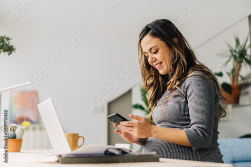Smiling woman expecting baby messaging on cellphone sitting at table with laptop