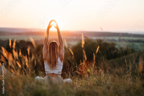 Back View on Woman Sitting in Meditation Yoga Pose and Catching Sun by Hands at Sunset Outdoors