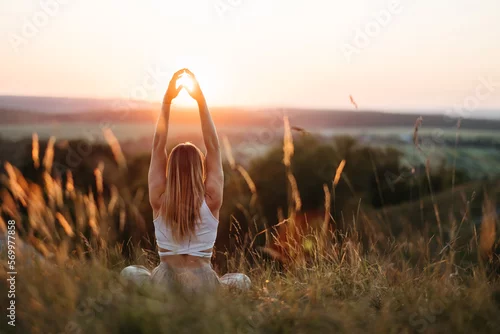 Obraz Back View on Woman Sitting in Meditation Yoga Pose and Catching Sun by Hands at Sunset Outdoors