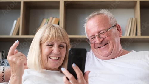 Smiling senior couple using smartphone browsing internet together sitting on sofa at home 