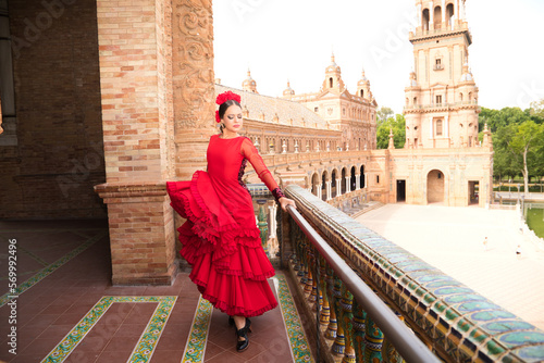 Beautiful teenage woman dancing flamenco on the balcony of a square in Seville. She wears a red dress with ruffles with a lot of art. Flamenco cultural heritage of humanity.