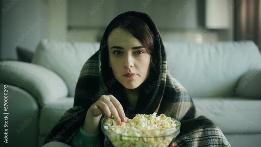 Camera view of addicted focused young woman watching interesting TV program film movie and eating popcorn alone at home Enchanted girl cannot take her eyes off the screen sitting on the floor indoors
