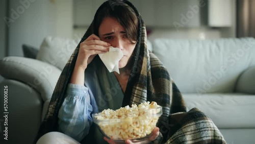 Camera view of sentimental young woman under the blanket at home alone and watch drama with popcorn Beautiful female with napkin wipes away tears after watching film or serial in the living room