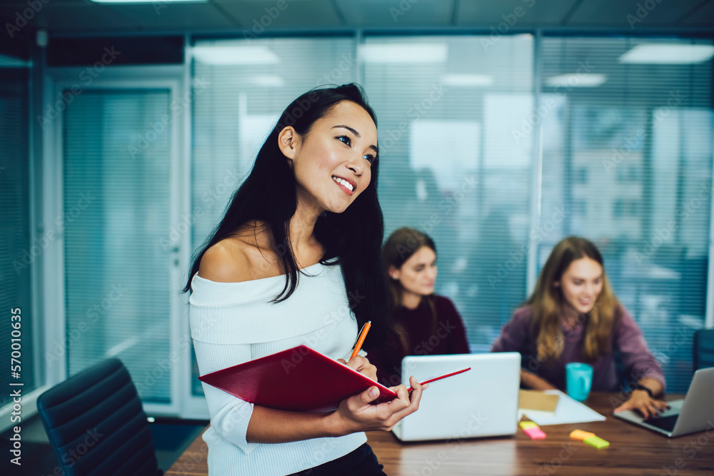 Thoughtful ethnic woman taking notes in planner near working colleagues Thoughtful ethnic woman taking notes in planner near working colleagues