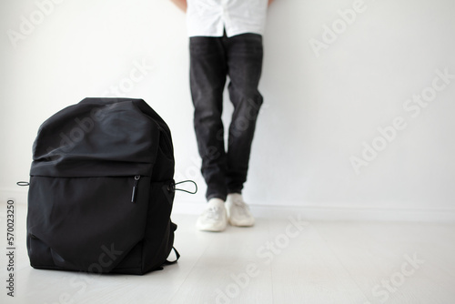 The guy with a bagpack on his shoulders is poaing on a white background. The teenager is wearing a white T-shirt and black jeans