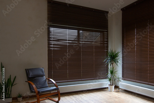 Wooden blinds on large windows in the interior. Living room with armchair and houseplants near windows with wood blinds. Motorized jalousie in the smart house. photo