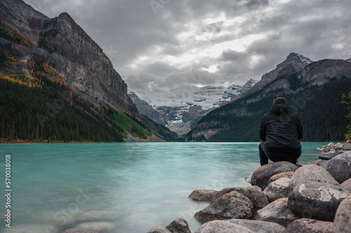 Young man sitting on her back enjoying the scenery of Lake Louise in Alberta, Canada, with crystal blue water and snow on the mountains in the background.