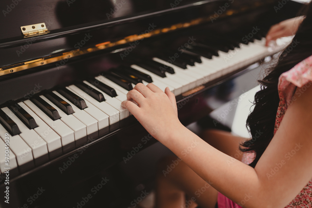 Fototapeta premium Cute and focused young little Asian girl practicing songs and melody with father and tutor while pressing white and black key and creating new music on piano as hobby and dedication at home