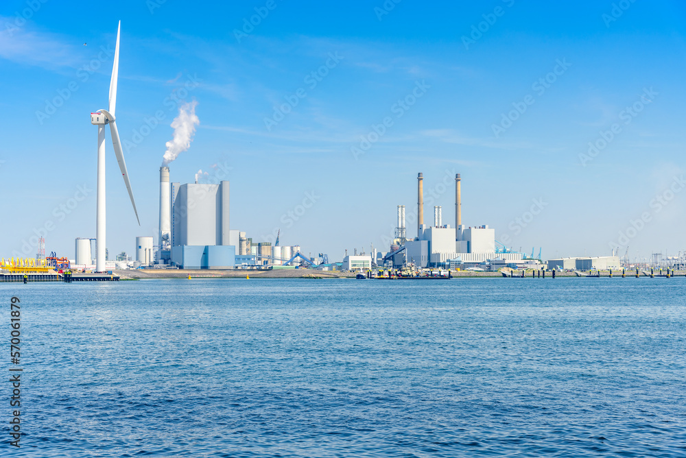 Naklejka premium Coal fired power plants with high smokestacks on a harbour. A tall wind turbine is in foreground.