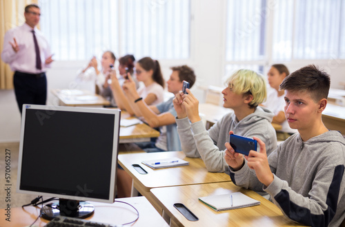 Group of modern teenagers sitting with mobile phones on lesson in classroom