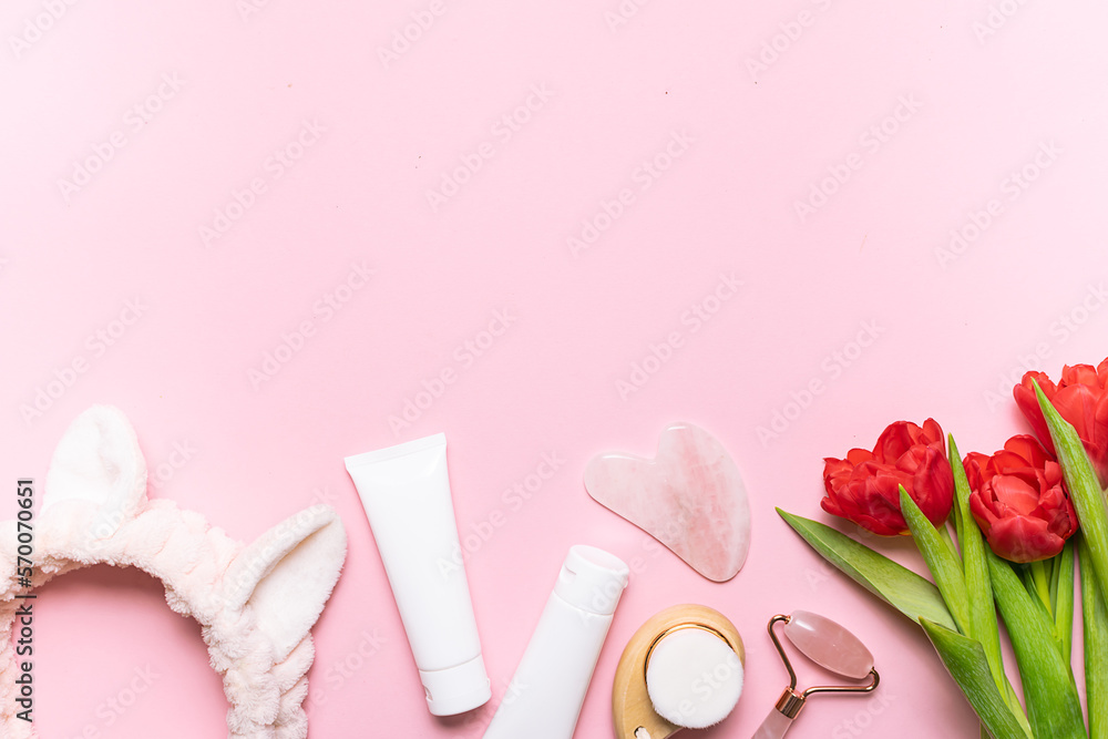 Face care concept. Asian Gua Sha massage tools, cream, mask in white plastic tubes, hair band and beautiful red tulips on light pink background with copy space for your design. Face care concept. Asian Gua Sha massage tools, cream, mask in white plastic tubes, hair band and beautiful red tulips on light pink background with copy space for your design.