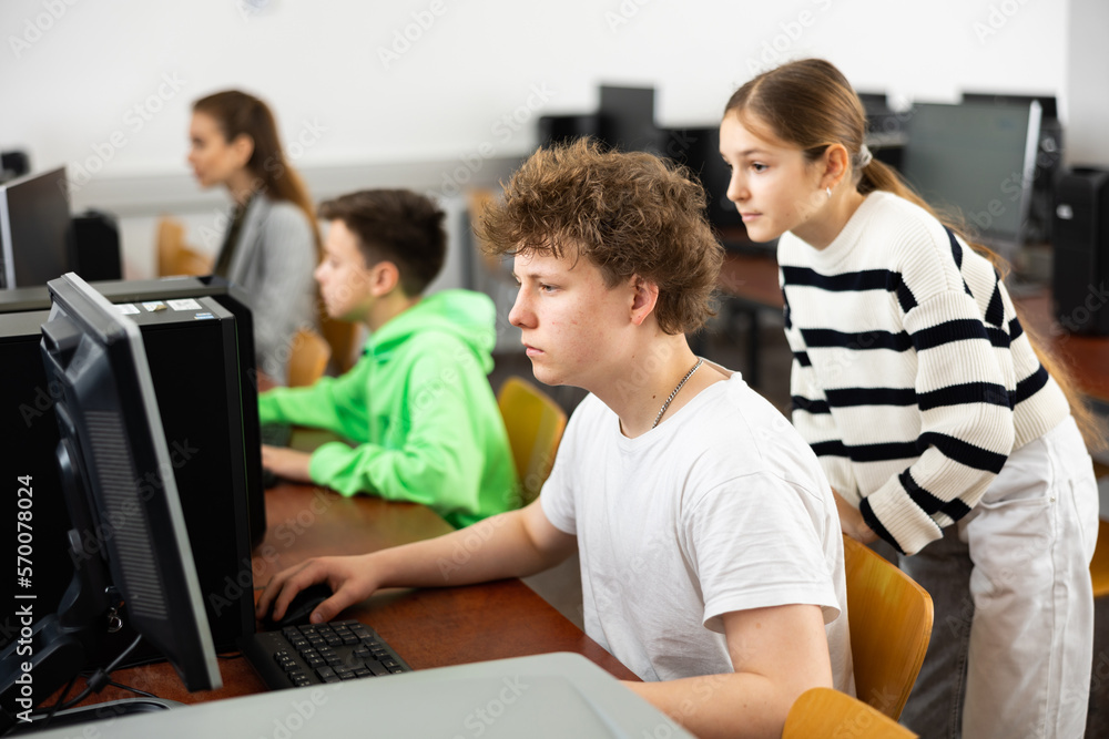 Teenage boy sitting at table and helping young girl with PC problem in computer class.