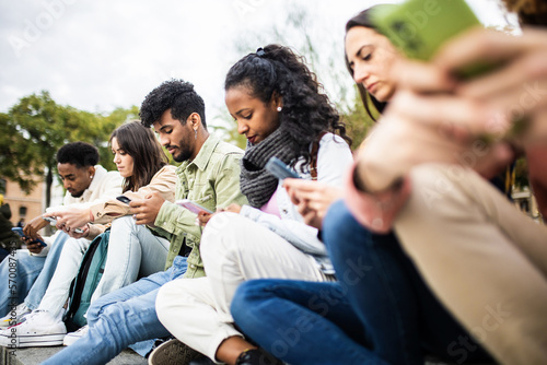 Addicted millennial generation people using mobile phone sitting outdoor. Young teenage group connected on smartphone app sharing social media content on