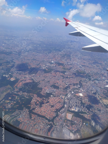White plane wing with red tip and city view from above with a blurred from the plane window and with the blue sky and clouds