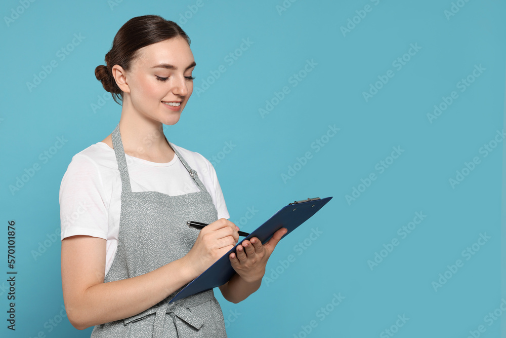 Beautiful young woman in clean apron with clipboard on light blue background. Space for text Beautiful young woman in clean apron with clipboard on light blue background. Space for text