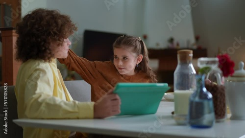 Rack focus from brother with digital tablet sitting at breakfast table to hyperactive sister playing laughing. Happy relaxed Caucasian boy and girl before school at home in the morning