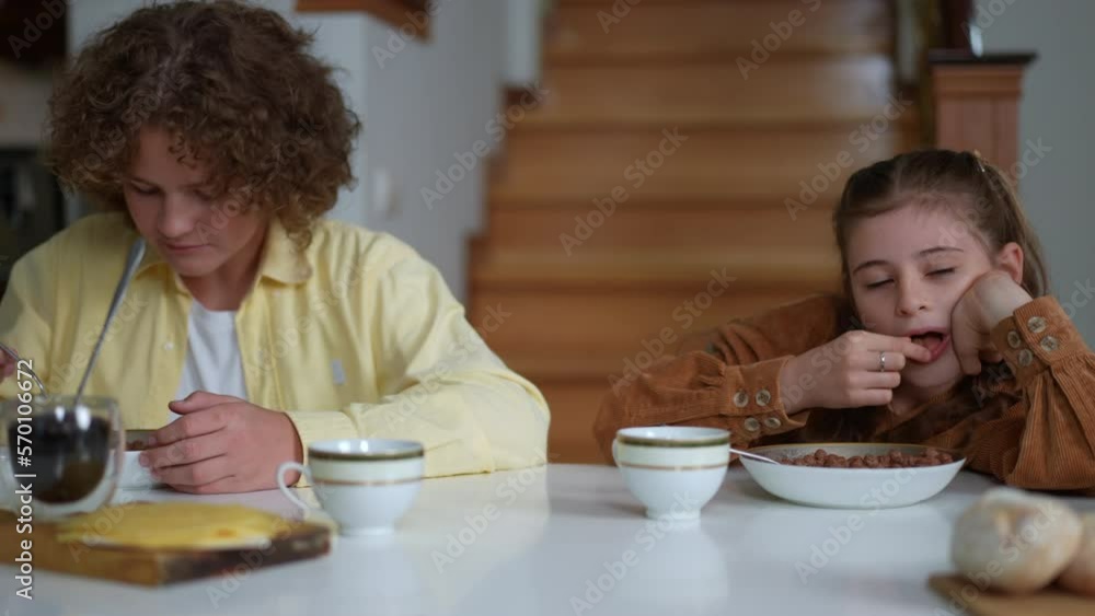 Front view portrait of schoolboy and schoolgirl eating morning cereal sitting at table in living room. Sleepy Caucasian girl putting head on table closing eyes as boy enjoying tasty breakfast Front view portrait of schoolboy and schoolgirl eating morning cereal sitting at table in living room. Sleepy Caucasian girl putting head on table closing eyes as boy enjoying tasty breakfast