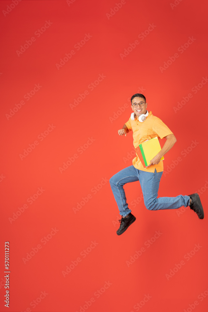 Young college man with glasses, jumping while looking at the camera, carrying his notebooks and headphones Young college man with glasses, jumping while looking at the camera, carrying his notebooks and headphones