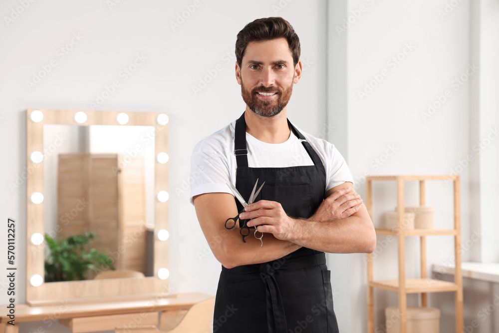Fototapeta premium Smiling hairdresser in apron holding scissors near vanity mirror in salon