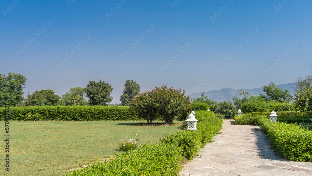Obraz premium A paved footpath runs through the park. White decorative lanterns are installed in the trimmed bushes on the roadsides. Trees and flowers grow on the lawn. Mountains against the blue sky. India