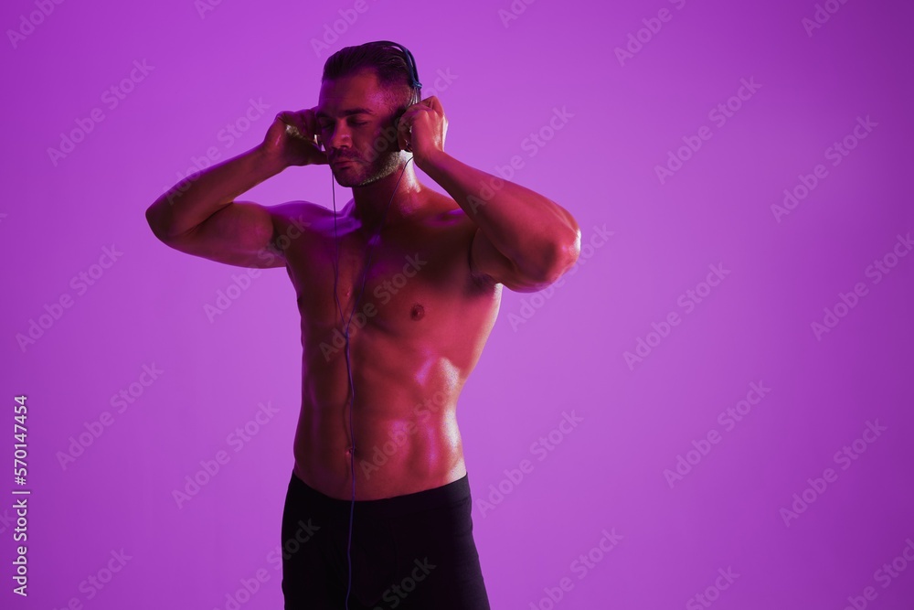 Man wearing headphones athletic physique bodybuilder nude torso abs full-length, fitness classes, purple colored light. Advertising, sports, active lifestyle, competition, challenge concept. 