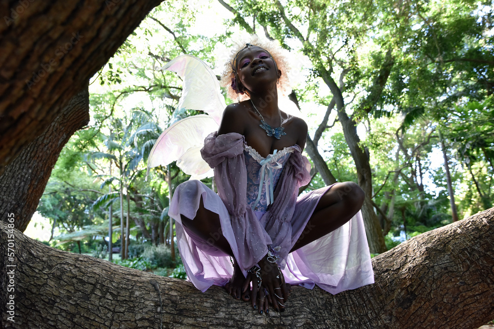 Portrait of beautiful African woman wearing purple fantasy costume, magical  fairy wings & flower crown afro, climbing tree branches in forest location  with natural glowing lighting. Stock Photo