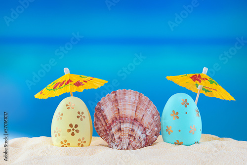 Happy easter. Yellow and blue eggs with conch shell under umbrellas on sandy beach of the sea or ocean. Holiday postcard in hot country. Travel, spring, summer. Copy space