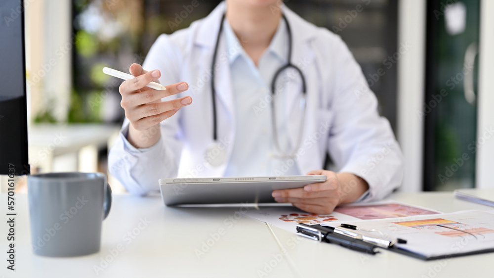 Fototapeta premium Cropped image of a professional Asian female doctor in uniform sits at her desk using tablet