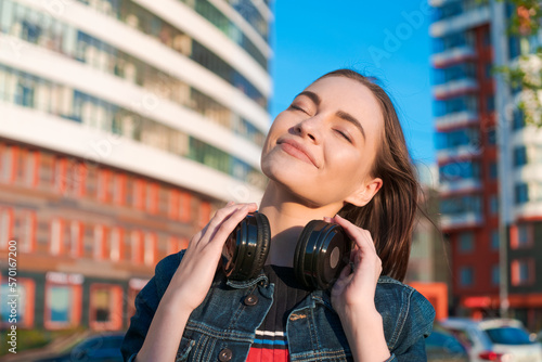 Young energetic cheerful caucasian woman wearing headphones. Dressed in a denim jacket, enjoying the spring sun. People urban youth lifestyle.