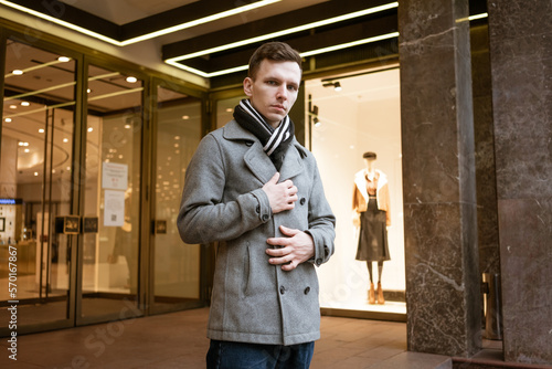 Beautiful model young guy in gray coat posing on city street during day. Caucasian man stylishly dressed