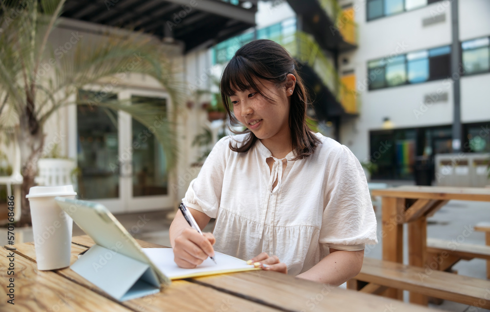 Fototapeta premium Young asian girl with freckles smiling and studying online with a tablet, notes and pen outdoors