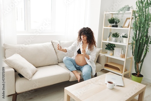Pregnant woman smile blogger showing baby feeding bottle filming herself on phone, video chat sitting on sofa at home freelancer in last month of pregnancy