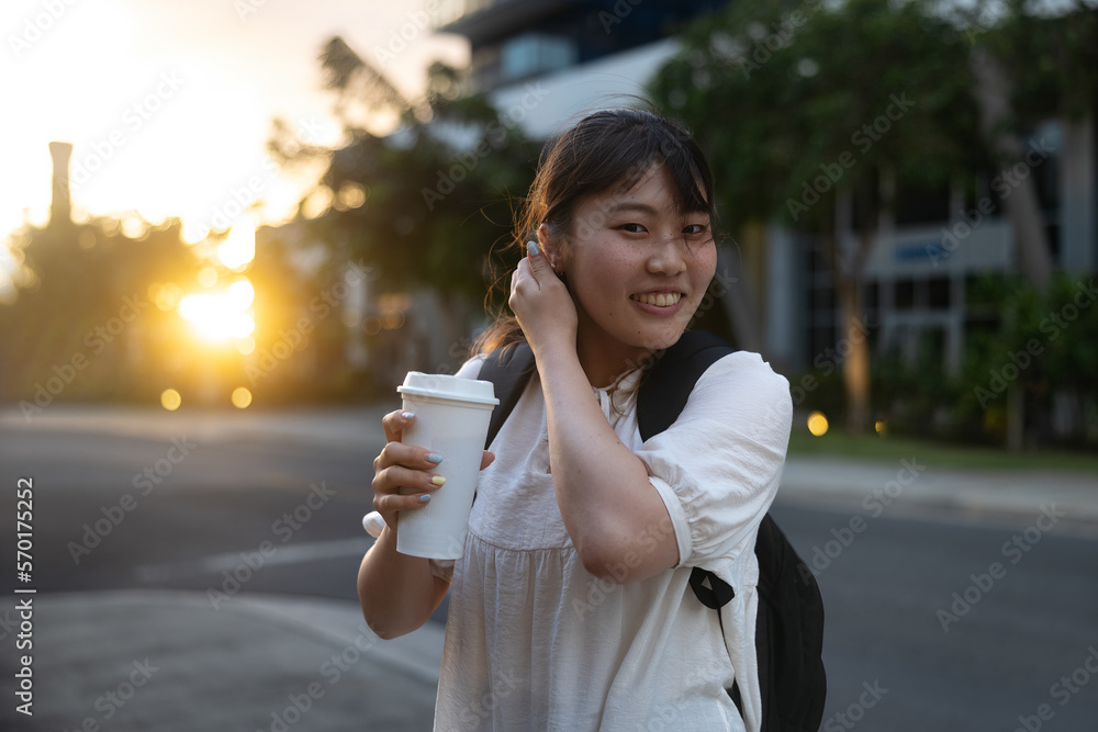 A young asian girl in a dress with a backpack smiling and walking on a street with a coffee or tea cup A young asian girl in a dress with a backpack smiling and walking on a street with a coffee or tea cup