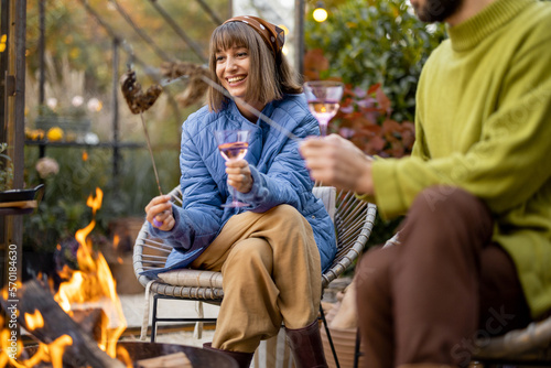Young stylish couple grilling food and warming up while sitting together by the fire, spending autumn evening time at cozy atmosphere in garden