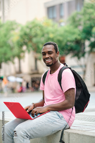 Man looking at camera and smiling while using a laptop outdoors.