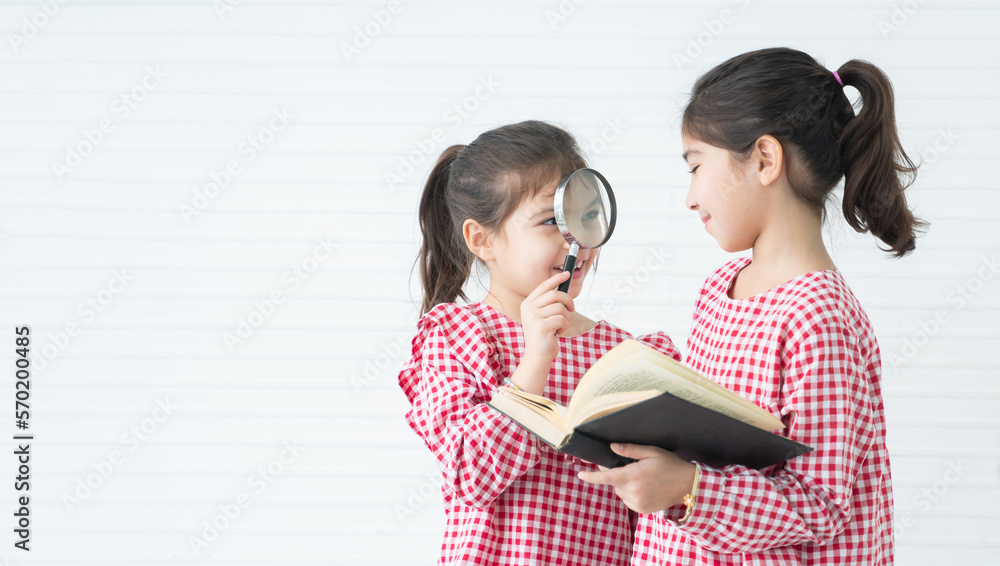 Two little cute children girls, reading a book, playing with magnifying glass while standing on white background. Siblings relationship and Education concept