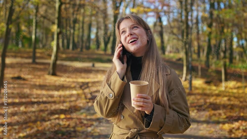 Side view of positive, young female walking in park with golden leaves. Beautiful lady wearing stylish clothes, talking on phone, drinking coffee. Concept of happiness and joy. Side view of positive, young female walking in park with golden leaves. Beautiful lady wearing stylish clothes, talking on phone, drinking coffee. Concept of happiness and joy.