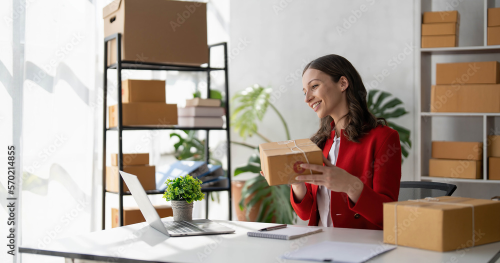 Happy young asian woman startup small business freelance holding parcel box and computer laptop and sitting on chair, Online marketing packing box delivery concept Happy young asian woman startup small business freelance holding parcel box and computer laptop and sitting on chair, Online marketing packing box delivery concept