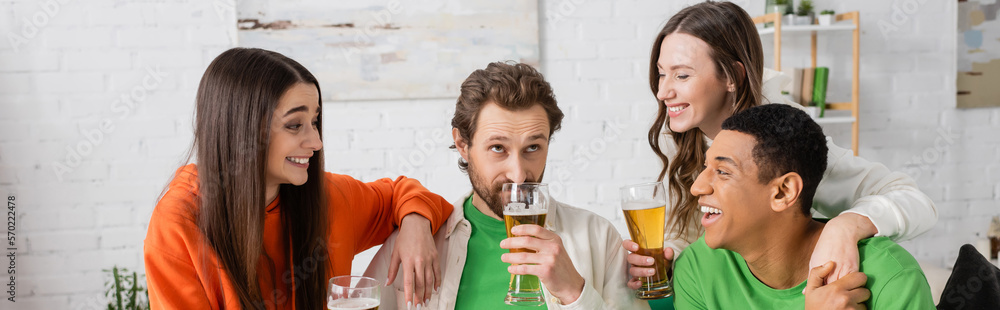 happy interracial group of people looking at bearded friend drinking beer in living room, banner