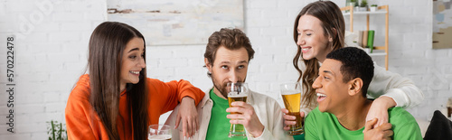 happy interracial group of people looking at bearded friend drinking beer in living room, banner