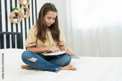 Child girl sitting and reading a book on bed in the living room at home. Kid girl reading a book in the library at the school