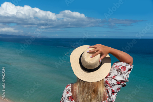 Mujer rubia de espaldas, con un vestido floreado, sosteniendo un sombrero de paja en una mano, mirando el mar turquesa de la costa de la turística Fuerteventura en las Islas Canarias