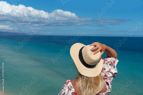 Mujer rubia de espaldas, con un vestido floreado, sosteniendo un sombrero de paja en una mano, mirando el mar turquesa y cristalino de la costa de la turística Fuerteventura en las islas Canarias