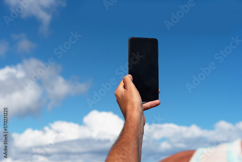 Primer plano del brazo de un hombre bronceado tomando una foto con su teléfono móvil del cielo azul con algunas nubes, durante un día soleado en las turísticas Islas Canarias. photo