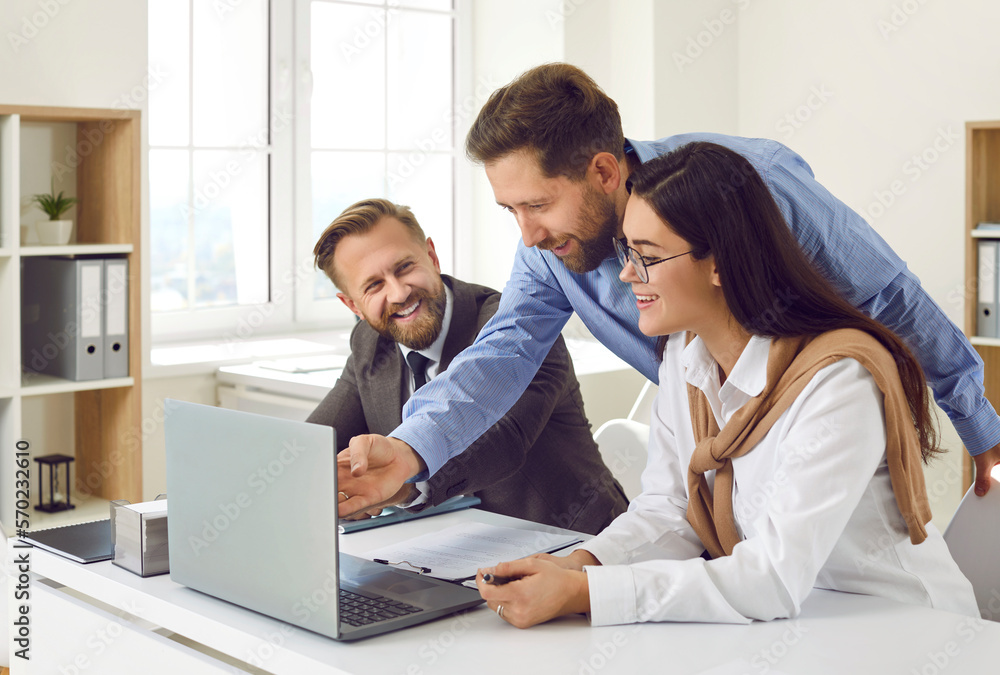 Work in office. Friendly cheerful colleagues in office together look at laptop screen and discuss business project. Man is showing something on computer screen to his smiling male and female coworkers Work in office. Friendly cheerful colleagues in office together look at laptop screen and discuss business project. Man is showing something on computer screen to his smiling male and female coworkers