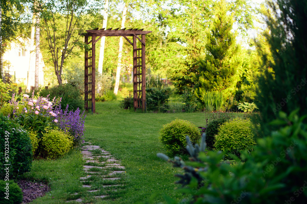 Beautiful summer garden view with curvy stone pathway and wooden archway. Natural woodland cottage garden with hostas, conifers and shrubs Beautiful summer garden view with curvy stone pathway and wooden archway. Natural woodland cottage garden with hostas, conifers and shrubs