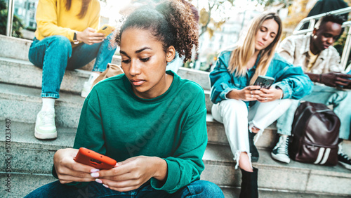 Multiracial young people using smart mobile phone device outside - University students looking smartphone sitting in college campus - Teenagers addicted to trendy technology and social media