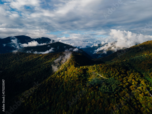 Drone landscape photo of the mountains in early Autumn. Some little clouds are flying low between mountains over it massive gray clouds and blue sky high. Sunlight breaking through the clouds.