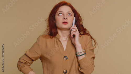 Redhead woman tired sleepy freelancer talking on mobile phone with friend, making online conversation. Disinterested sad young girl having annoyed boring talk on smartphone on beige studio background